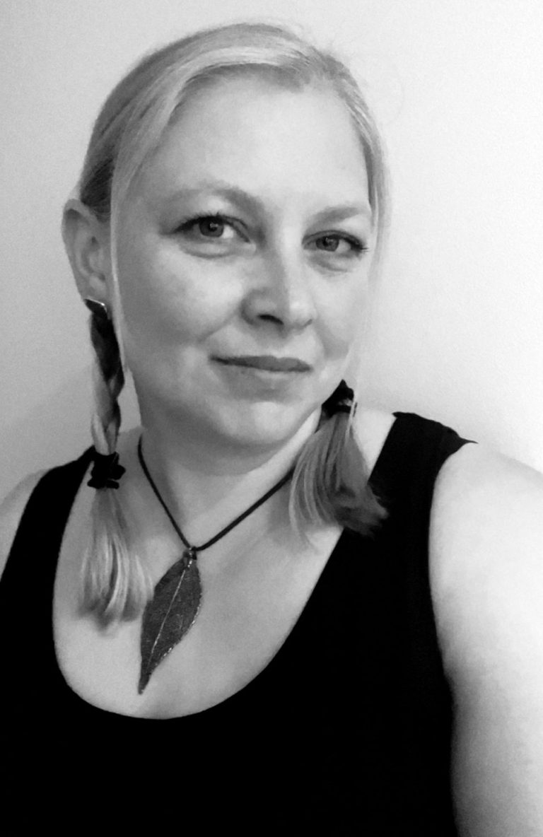 Portrait of a Tesi Hughes with long hair, wearing a black top and necklace, set against a plain background.